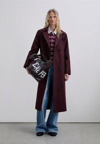 Persona vestida con un abrigo trench burdeos, camisa a rayas, jeans azules, sosteniendo un gran bolso de mano negro con el texto blanco "LOCAL PER".