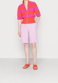 Persona che indossa un maglione con zip a strisce rosse e rosa, pantaloni corti viola chiaro fino al ginocchio e sandali con tacco aperti rossi, in piedi davanti a uno sfondo neutro.