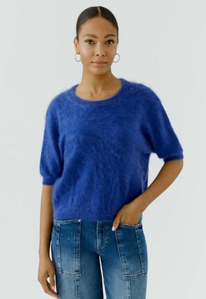Jonge vrouw met haar naar achteren gebonden draagt gouden oorringen, een pluizige blauwe korte mouwen trui en een high-waisted spijkerbroek met voorzakken.