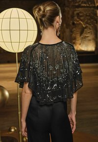 Must have kvaliteetne must läbipaistev topp, millel on sädelev lillemuster, lainelised varrukad ja ümmargune kaelus; kombineeritud libedate mustade pükstega. Riietuse tagumine vaade.