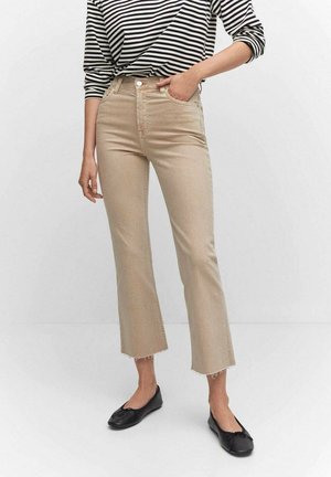Persoon draagt beige high-waist cropped broek met ruwe zomen, gestreept shirt met lange mouwen en zwarte ballerina's, staat met één hand in de zak.