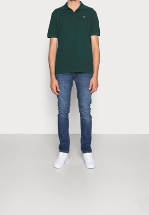 Mand iført mørkegrøn polo, blå slim-fit jeans og hvide sneakers, stående mod en ensartet lys grå baggrund.