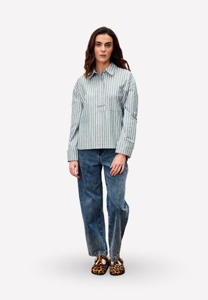 Gestreepte button-up shirt in groen en wit, gecombineerd met losse blauwe spijkerbroeken en schoenen met dierenprint, gedragen door een model.