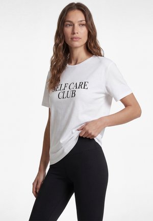 Frau mit langen braunen Haaren, die ein weißes T-Shirt mit dem Text "SELF CARE CLUB" und schwarze Leggings trägt, steht vor einem einfarbigen Hintergrund.