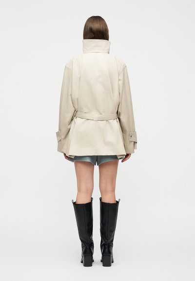 Άτομο που φοράει μπεζ μπουφάν με ζώνη και ψηλό γιακά, ανοιχτό μπλε σορτς και μαύρες μπότες με τακούνια μέχρι το γόνατο, στέκεται με την πλάτη γυρισμένη σε λευκό φόντο.