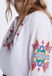 Blouse blanche brodée présentant des motifs floraux et géométriques vibrants en rouge, bleu, jaune et vert sur les manches et le col.