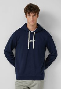 Giovane in piedi con le mani dietro la schiena, indossa una felpa blu navy con cordini bianchi e pantaloni grigio chiaro su uno sfondo semplice.