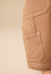 Close-up of beige fabric pocket with detailed stitching on garment against neutral background.
