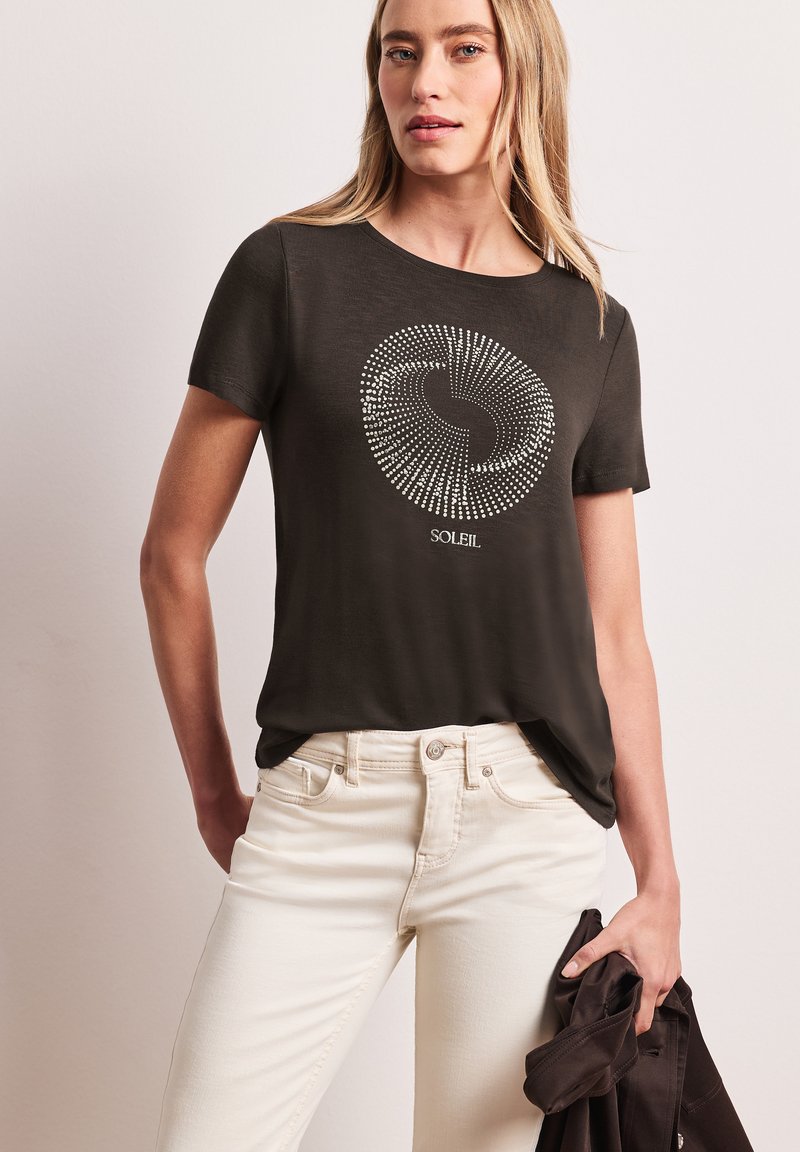 Donna con maglietta nera con design circolare a puntini e la parola "SOLEIL", che tiene una giacca scura piegata, abbinata a pantaloni bianchi.