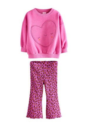 Junges Mädchen steht drinnen und trägt ein leuchtend pinkes Sweatshirt mit einem lächelnden Herz und rosa Leopardenhose.