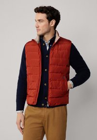 Chaleco acolchado rojo con cuello alzado, combinado con un suéter azul marino. Presenta forro beige y un bolsillo lateral, llevado con pantalones beige.