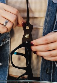 Mani con smalto rosso scuro che attaccano occhiali neri a una cinghia in pelle marrone sul collo, sopra una maglietta beige a coste e una giacca di jeans.