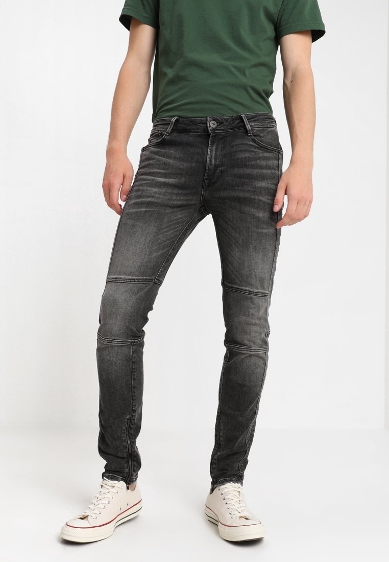 Vaqueros de denim negros con un ajuste slim, que presentan un ligero desgaste y detalles de costura, combinados con una camiseta verde y zapatillas blancas.