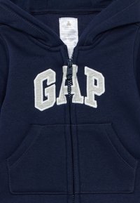 Σκούρο μπλε φλις φούτερ με φερμουάρ και τσέπη καγκουρό, με λευκό κεντητό λογότυπο «GAP» στο μπροστινό μέρος.