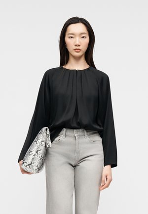 Žena v volném černém topu s dlouhým rukávem a světle šedých džínách, držící bílou a černou psaníčko s hadím vzorem.