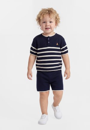 Νήπιο με ξανθά, σγουρά μαλλιά που φοράει μπλε-λευκή ριγέ μπλούζα, σκούρες μπλε βερμούδες και λευκά αθλητικά παπούτσια, περπατάει προς τα εμπρός.