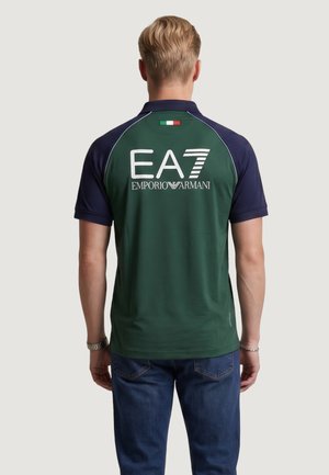 Mężczyzna noszący ciemnozieloną i granatową koszulkę polo z logo „EA7 Emporio Armani” i małą włoską flagą na górnej części pleców, połączoną z niebieskimi dżinsami.