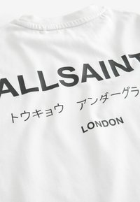 Valge puuvillane T-särk musta tekstiga seljal, kus on kirjas "ALLSAINTS", koos täiendavate sümbolitega ja "LONDON" allosas.
