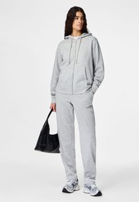 Mujer con sudadera con capucha gris claro con cremallera y pantalones deportivos a juego, sosteniendo un bolso negro y calzando zapatillas blancas y grises.