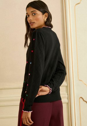 Černý pletený svetr se vzorem diamantů, zdobený barevnou výšivkou srdcí na rukávech a s volánkovým růžovým lemem na manžetách a kolem výstřihu.