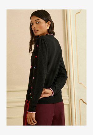 Melns adīts džemperis ar dimanta rakstu, uz piedurknēm krāsaina sirds izšuvuma un rozā volānu apdare aprocēs un apkaklē.