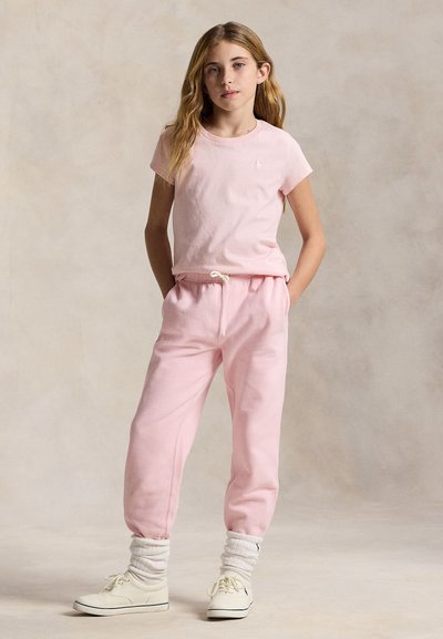 Fată stând cu mâinile în buzunare, purtând un tricou roz pal, pantaloni de trening asortați, șosete albe adunate și adidași albi.