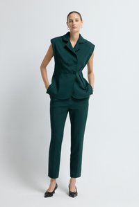 Gilet verde su misura senza maniche con doppi bottoni, abbinato a pantaloni aderenti dello stesso colore e scarpe nere con tacco basso.