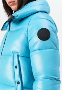 Close-up van een persoon die een glanzende lichtblauwe pufferjas met capuchon draagt, met een zwart ronde "Save the Duck" logo patch op de mouw.