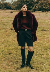 Burgundijas krāsas mākslīgā kažokāda, tekstūrēts burgundijas svīteris, melni šorti ar zelta jostu un melnas augstās zābaki uz zālāja.