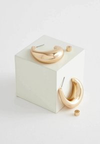 Guldtonade ringörhängen med en slät, glansig yta. Distinkt böjd form, placerade på en vit kub med en liten cylindrisk accentdel.