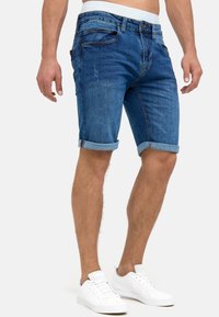 Shorts in denim di un blu medio con orli arrotolati, design a cinque tasche e chiusura con bottone. Abbinati a sneakers bianche.
