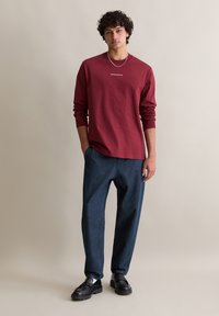 Rotes Langarmshirt mit Rundhalsausschnitt und Logo, kombiniert mit locker sitzenden dunklen Jeanshosen und schwarzen Slippern. Sanfte Texturen, lässiges Design.