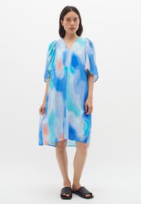 Blauwe en pastelgekleurde jurk met korte pofmouwen, V-hals, vloeiende stijl, knie-lengte, gemaakt van lichte stof. Zwarte instap schoenen.