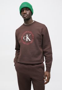 Barna pulóver kör alakú logóval, amelyen piros és fehér betűkkel szerepel a "CALVIN KLEIN NEW YORK SINCE 1968" felirat. Hozzáillő barna nadrággal és zöld sapkával párosítva.