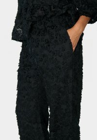 Czarne spodnie o fakturze z kwiatowymi wzorami, ręka swobodnie umieszczona w bocznej kieszeni, noszone z dopasowaną, fakturowaną bluzką.