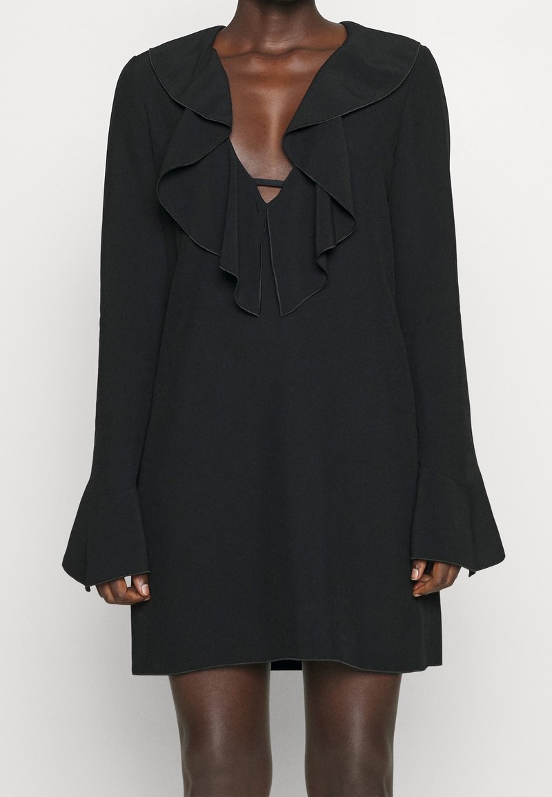 Robe noire à manches longues avec encolure en V à volants et poignets évasés portée par une personne à la peau foncée, debout devant un fond uni.