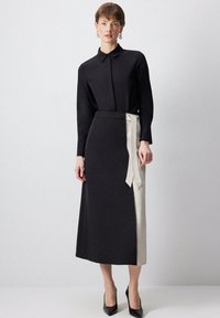 Robe noire à manches longues avec un col, taille cintrée et détail en ruban beige sur le côté ; accompagnée de talons hauts noirs.