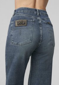 Jeans dżinsowe w ciemnym niebieskim kolorze, z teksturowanym wykończeniem, standardowymi tylnymi kieszeniami oraz naszywką z logo w pobliżu pasa.