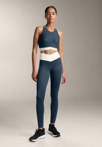 Atletski komplet v temno zeleni barvi, ki vključuje crop top z belimi poudarki in hlače z visokim pasom. Hlače so elegantne in s gladko teksturo.