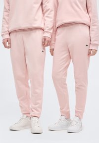 Δύο άτομα που φορούν ταιριαστά ανοιχτό ροζ φόρμες και φούτερ, συνδυασμένα με λευκά αθλητικά παπούτσια, στέκονται δίπλα δίπλα.