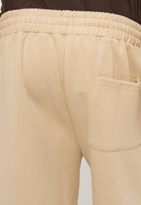 Μπεζ παντελόνι με ελαστική μέση και μία πίσω τσέπη, εμφανιζόμενο από τη μέση έως το μέσο του μηρού, φορεμένο με σκούρο τοπ.