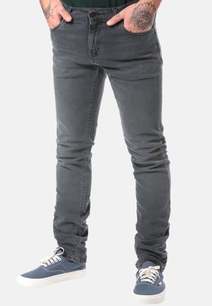 Mann trägt ausgewaschene schwarze Skinny-Jeans mit Händen in den Taschen und blaue Canvas-Sneakers mit weißen Schnürsenkeln und Sohlen.