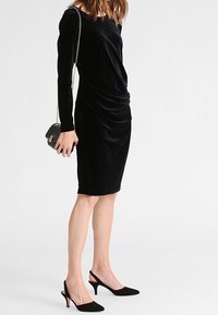 Robe longue à manches longues en velours noir avec un design drapé, longueur au genou, associée à des escarpins noirs à bouts pointus et une petite sacoche épaule noire.