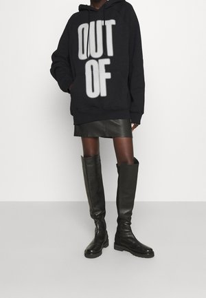 Henkilö pukeutuneena mustaan yläosalta väljään huppariin, jossa on teksti "OUT OF", mustaan nahkahameeseen ja polven yli ulottuviin mustiin nahkasaappaisiin.