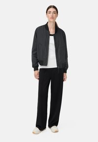 Veste bomber noire à texture lisse, poignets côtelés et fermeture éclair avant, associée à un haut blanc et un pantalon large noir. Chaussures neutres.