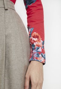 Pitkähihainen punainen paita, jossa on vaaleanpunaisia, sinisiä ja oransseja kukkakuoseja, yhdistettynä vaaleanruskeaan, teksturoituun korkeavyötäröiseen hameeseen.