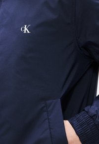 Μπλε σκούρο μπουφάν κατασκευασμένο από ελαφρύ, υφασμάτινο υλικό με υφή. Διαθέτει ελαστικές μανσέτες, πλαϊνή τσέπη και λευκό λογότυπο στο στήθος.