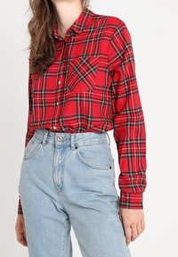 Kvinde iført en rød ternet knap-up skjorte stoppet ned i højttaljede lyseblå jeans, stående mod en ensartet baggrund.