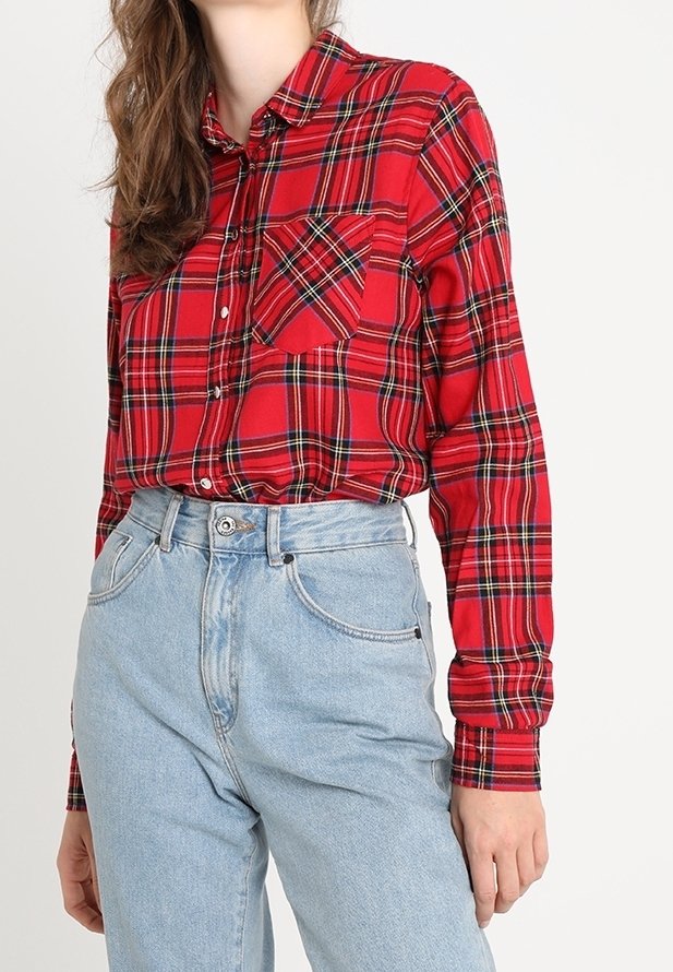 Kvinde iført en rød ternet knap-up skjorte stoppet ned i højttaljede lyseblå jeans, stående mod en ensartet baggrund.