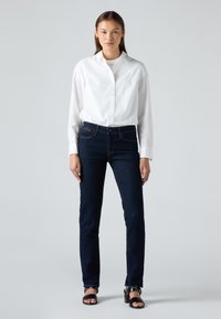 Γυναίκα που στέκεται φορώντας ένα λευκό πουκάμισο με κουμπιά, σκούρο μπλε στενό τζιν και μαύρες ανοιχτές ψηλοτάκουνες σαμπάνες μπροστά σε απλό γκρι φόντο.